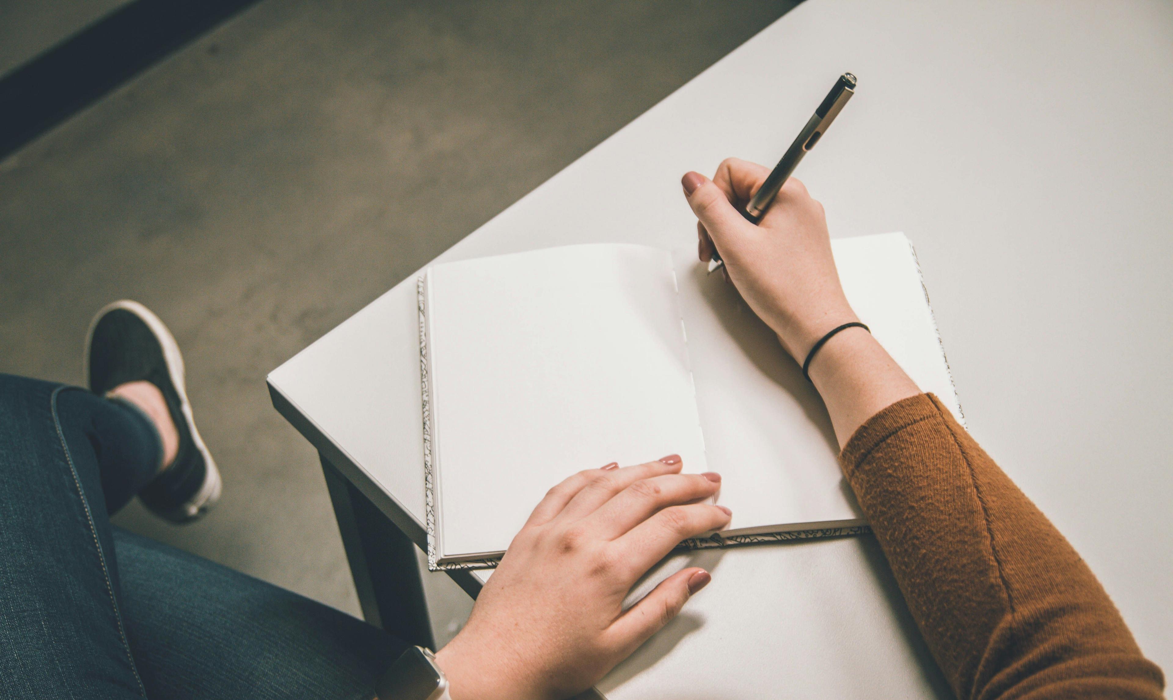 A close-up of a person's hand writing in an open notebook with a pen, sitting at a desk, representing brainstorming or planning tasks.