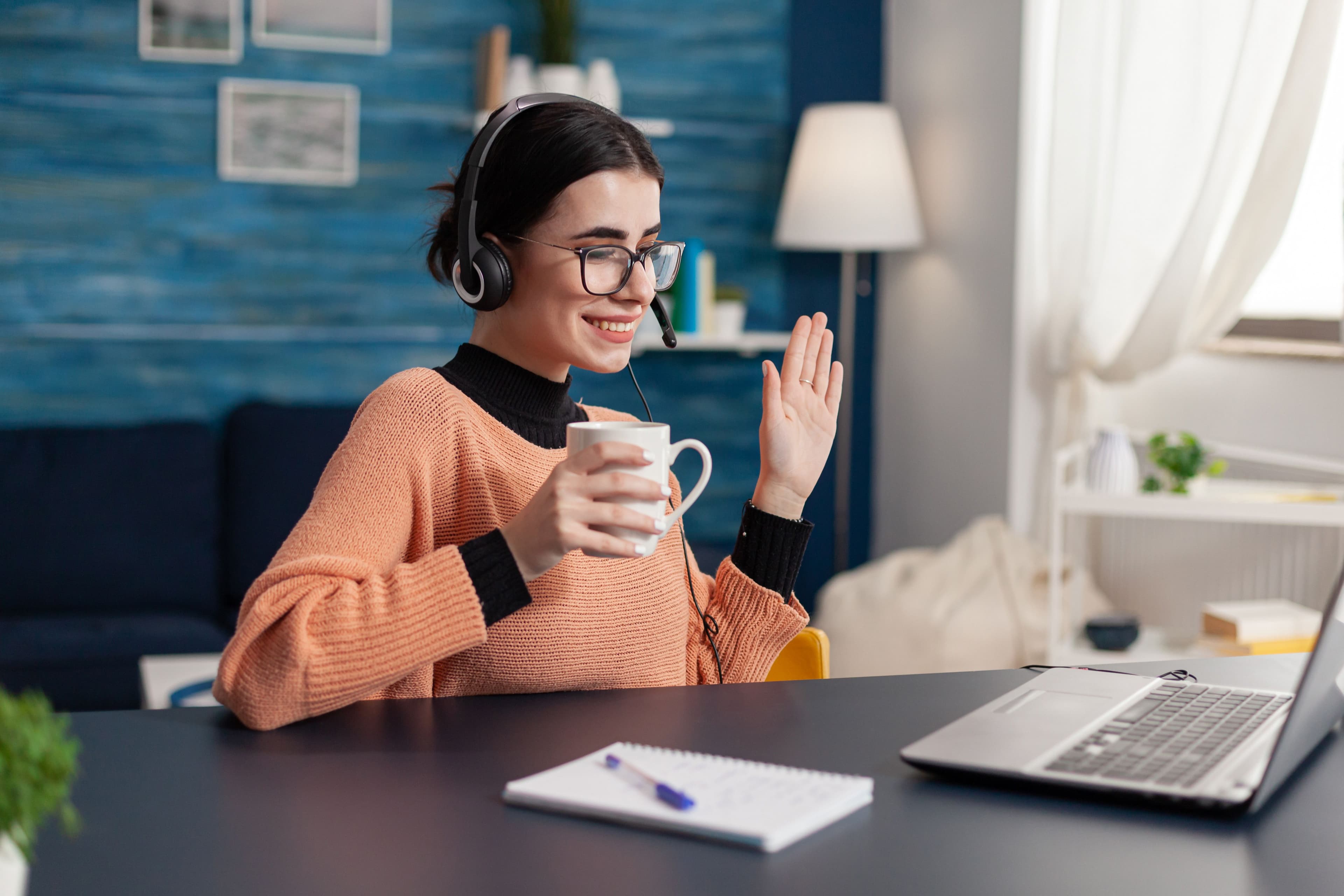 A woman in a cozy sweater and glasses smiling while waving during a video call, holding a coffee mug. She is at a desk with a laptop and a notepad. Perfect for small business websites promoting virtual customer service or online consultations.