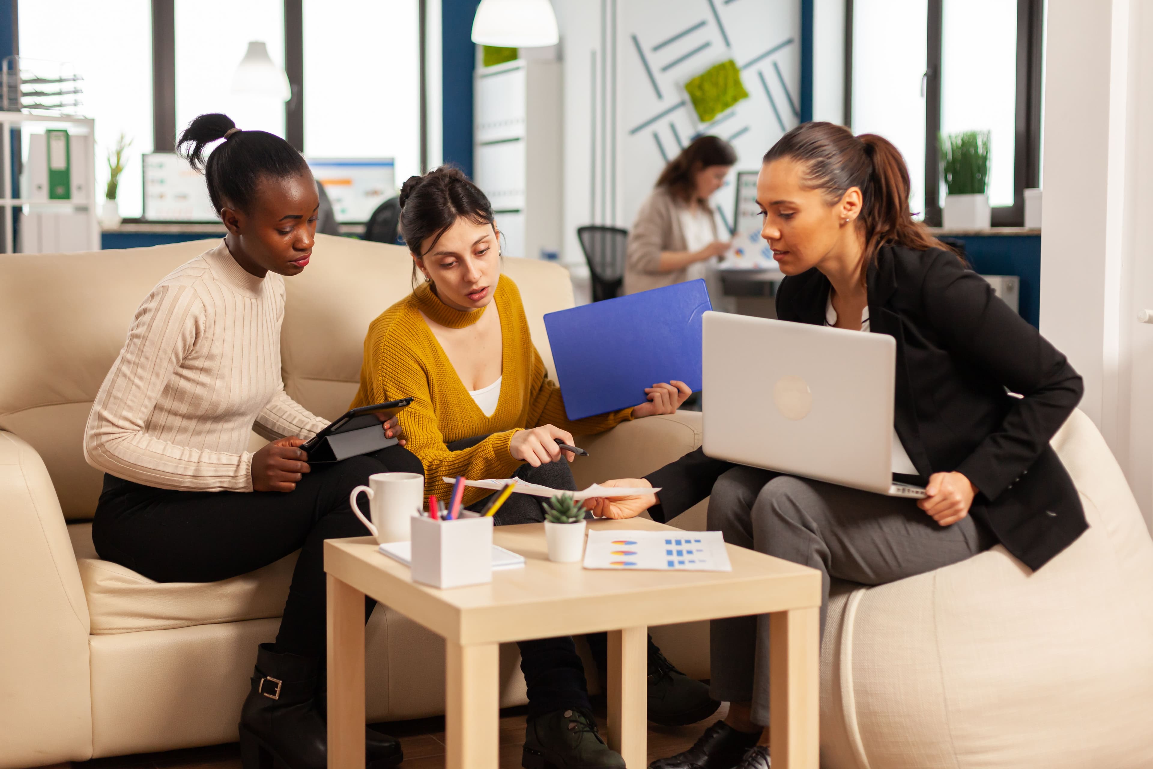 Three diverse businesswomen engaged in a meeting, discussing stock market performance reports, with laptops and documents in a cozy office lounge area.