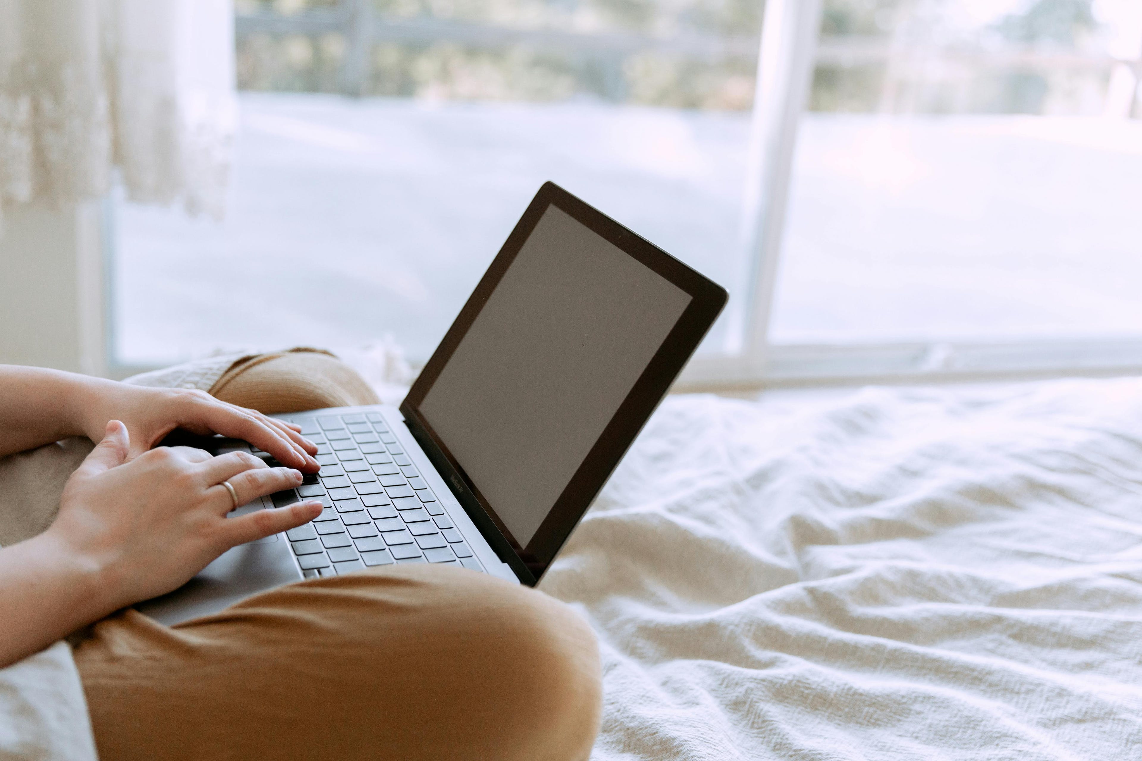A person is typing on a laptop, comfortably sitting on a bed with a light blanket, focusing on marketing and analytics tasks in a serene, natural light setting.
