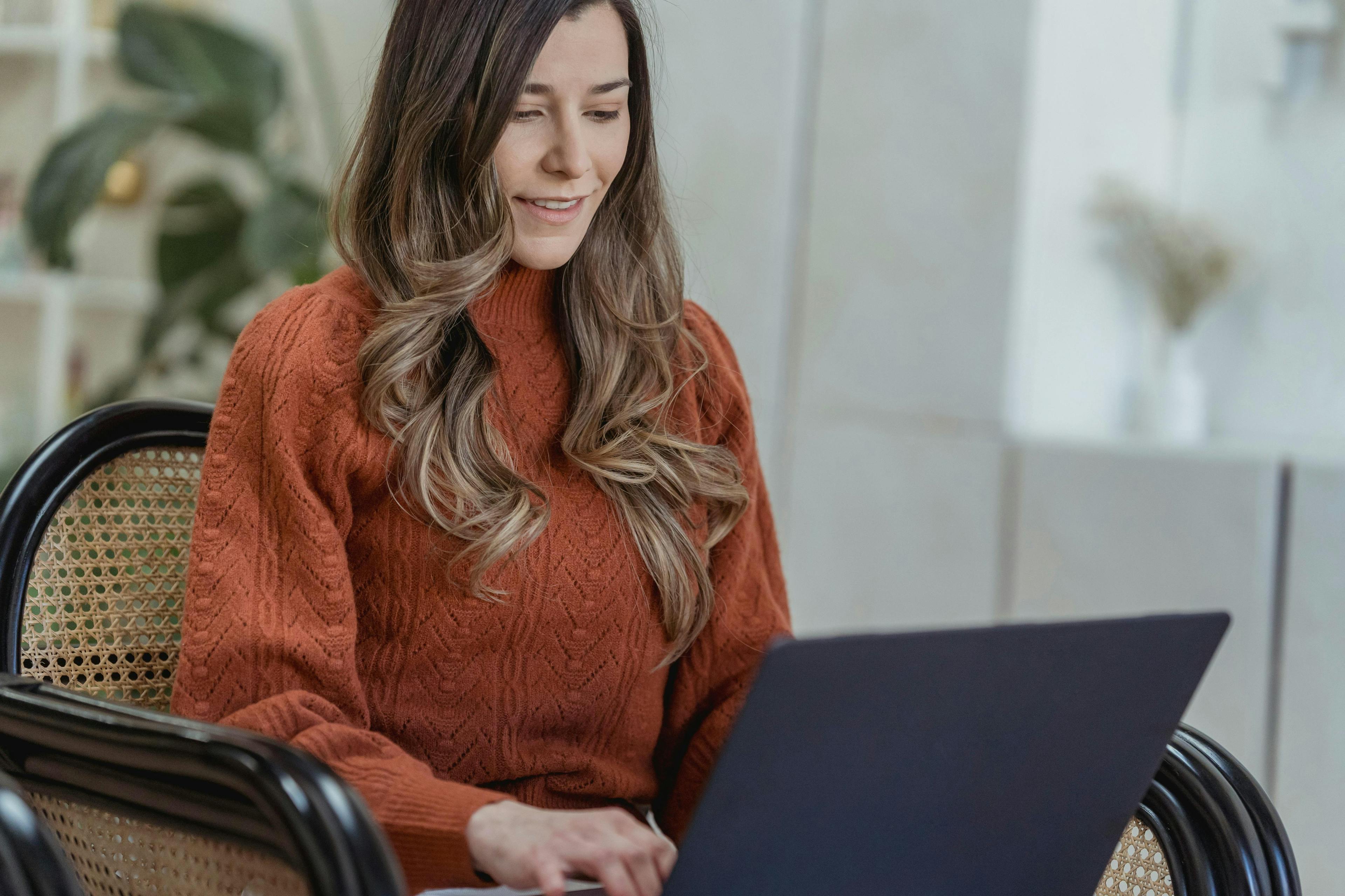 a woman sitting on a chair, smiling as she works on a laptop. The cozy and welcoming indoor setting highlights a conducive atmosphere for connecting with "lead generation companies" to enhance marketing strategies and business growth.