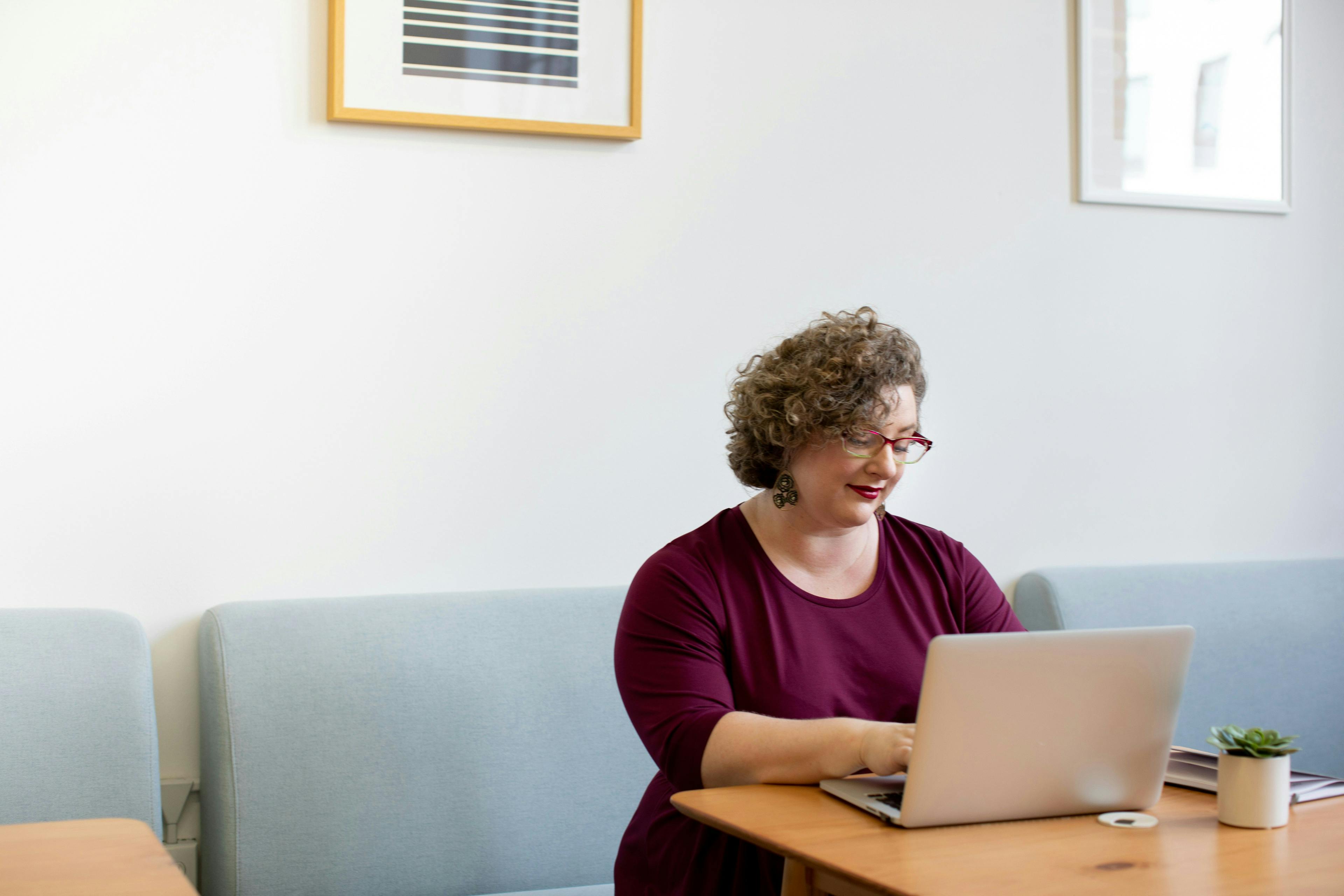 A woman with curly hair and glasses working on a laptop at a wooden table in a modern, minimalist room. The text 'how to create a landing page on Shopify' is implied by the context.